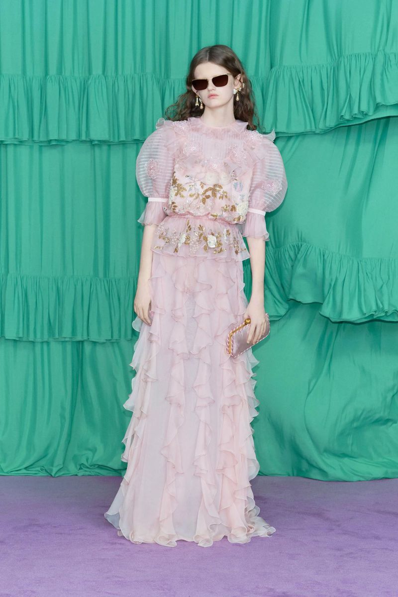Full-length shot of a woman with long hair, wearing sunglasses, a pink layered dress and holding a clutch, posing in front of a teal backdrop.