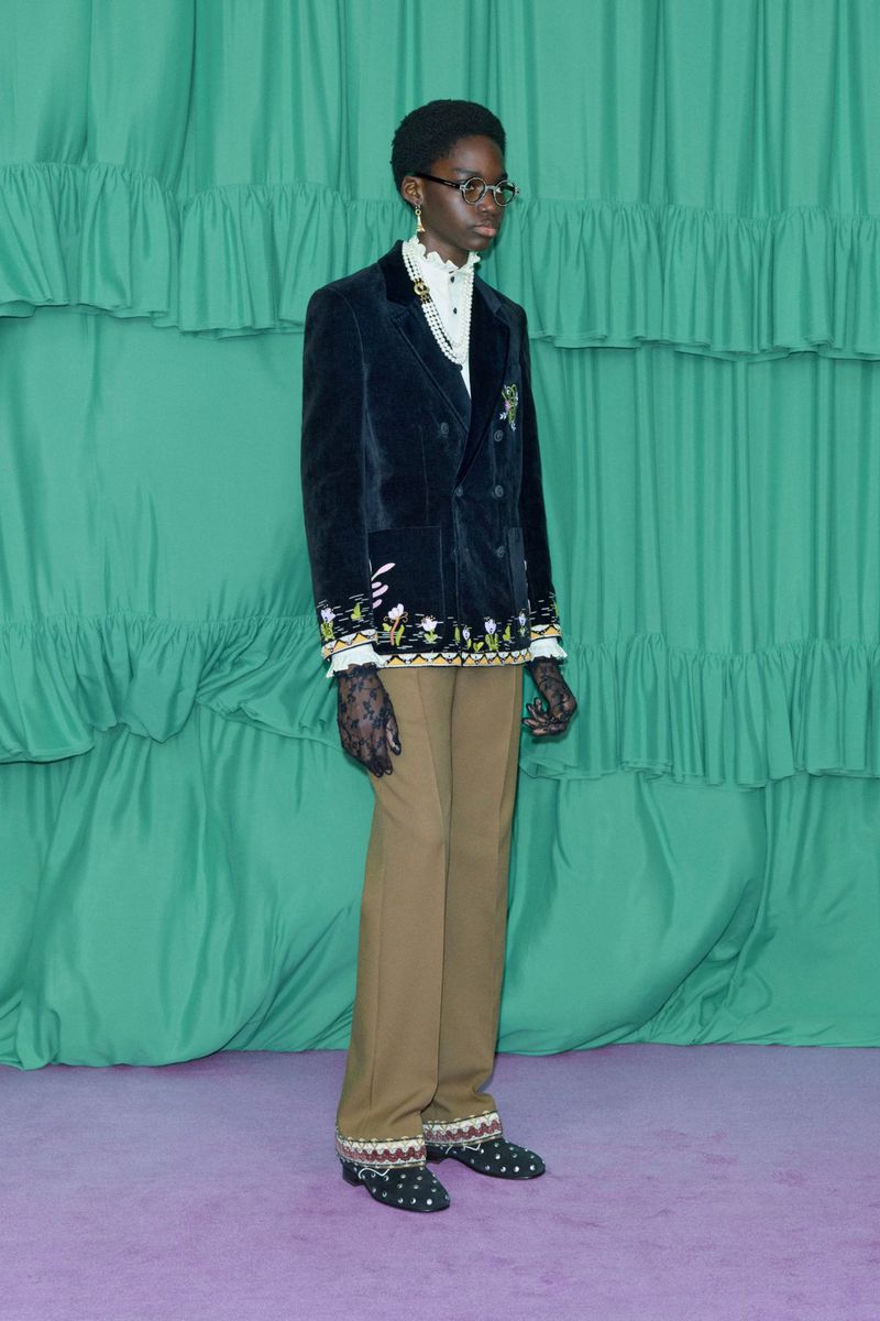 A person in a black embroidered velvet blazer and tan trousers stands against a green backdrop with ruffles, showcasing a formal fashion look.