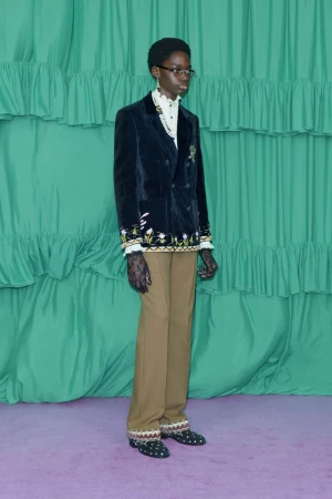 A person in a black embroidered velvet blazer and tan trousers stands against a green backdrop with ruffles, showcasing a formal fashion look.