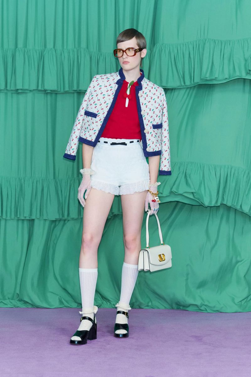 A woman models a white jacket with blue trim and cherry pattern with white shorts, socks, heels, and a white purse against a green backdrop.
