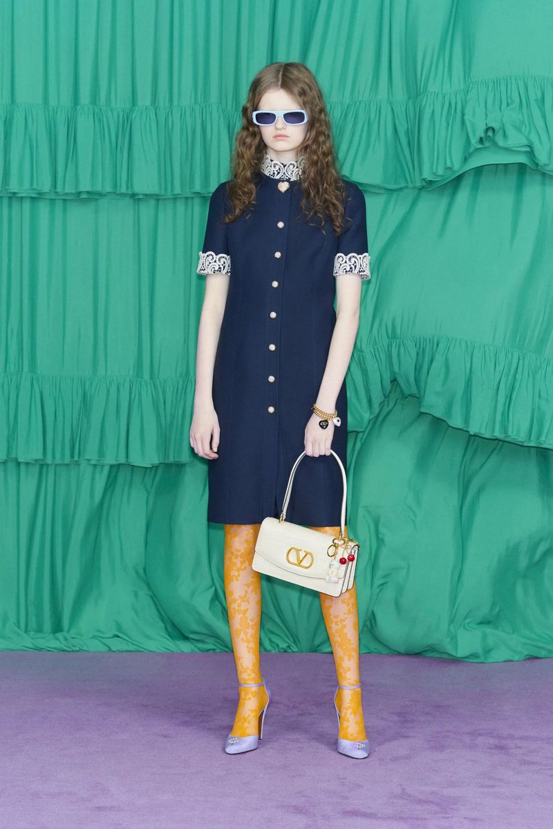 Adult female fashion model poses in knee-length navy dress with lace trim, sunglasses, and a cream-colored purse against a green backdrop.