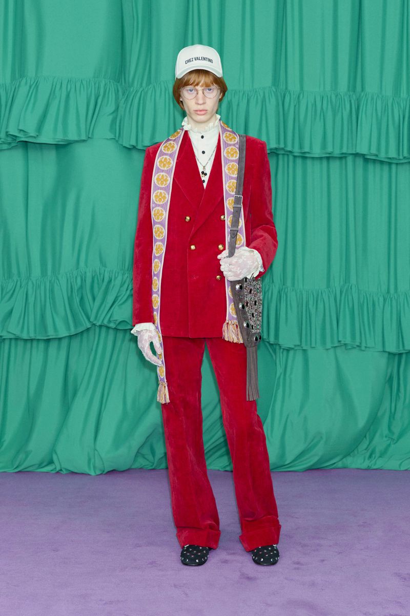Red-suited person with cap and shoulder bag stands against a ruffled teal backdrop, creating a bold fashion statement.