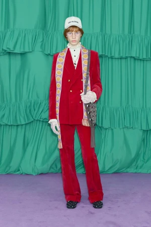 Red-suited person with cap and shoulder bag stands against a ruffled teal backdrop, creating a bold fashion statement.