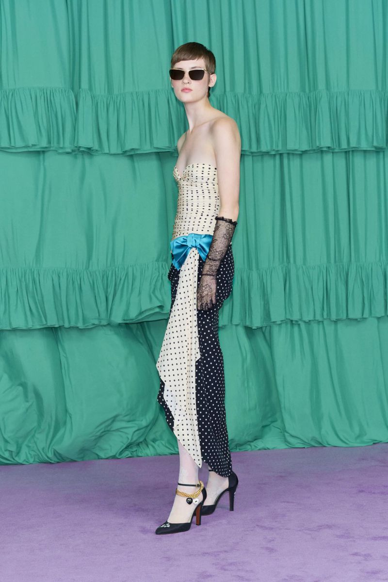 A fashion model poses in a strapless dotted top and polka dot pants in front of a ruched green curtain backdrop in a studio.