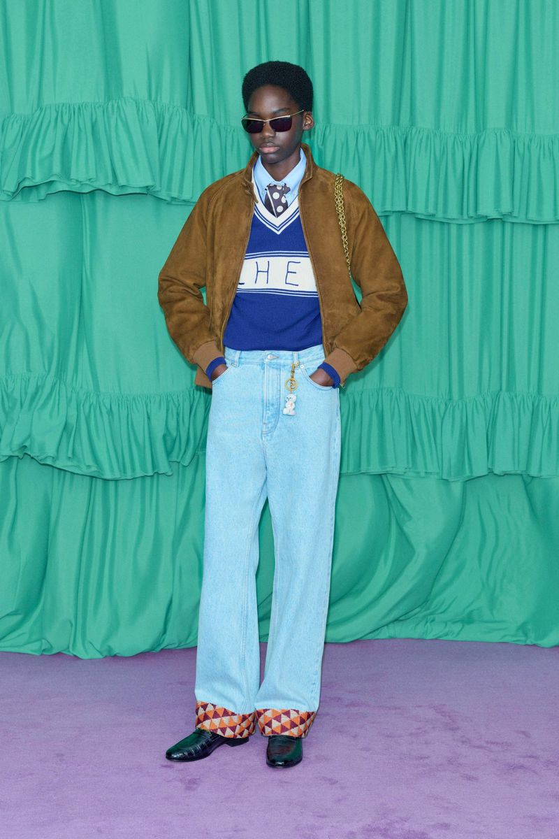 A person wearing a brown jacket, blue sweater, and light blue jeans poses in front of a teal backdrop with horizontal ruffles.