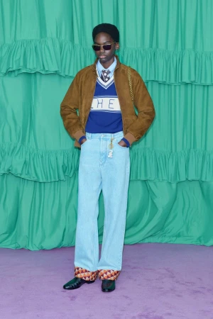 A person wearing a brown jacket, blue sweater, and light blue jeans poses in front of a teal backdrop with horizontal ruffles.