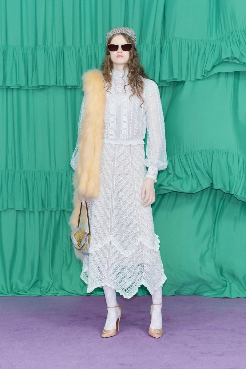 A slender woman in a long-sleeved lace dress, sunglasses, and heels stands in a studio with a green ruffled backdrop.