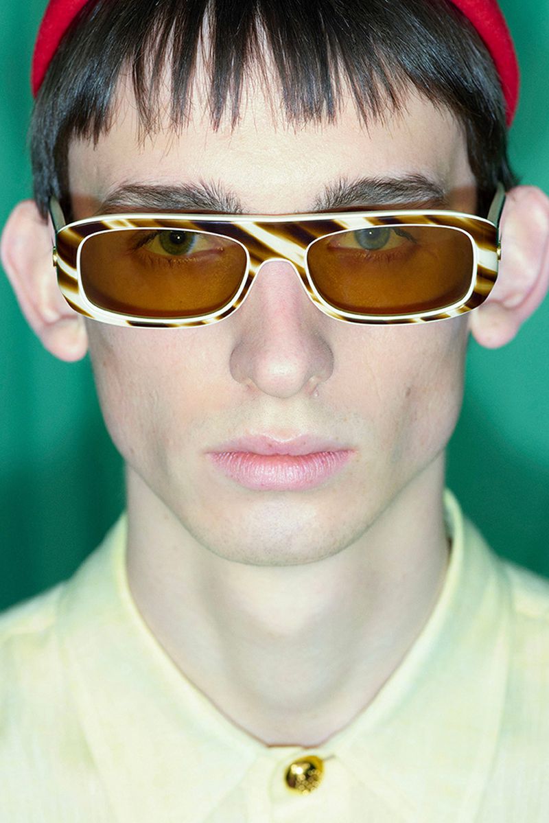 Close-up studio shot of an adult male wearing yellow shirt, red hat, and striped sunglasses against a green backdrop, fashion headshot.