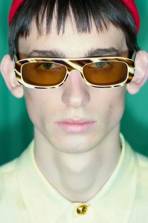 Close-up studio shot of an adult male wearing yellow shirt, red hat, and striped sunglasses against a green backdrop, fashion headshot.