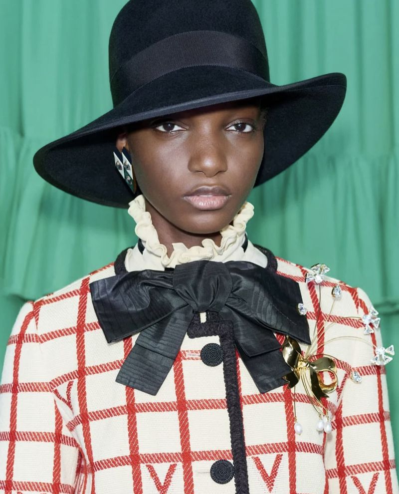 Portrait of an African woman wearing a wide-brim hat and red-and-white plaid jacket with a large bow, accessorized with statement earrings and a brooch.
