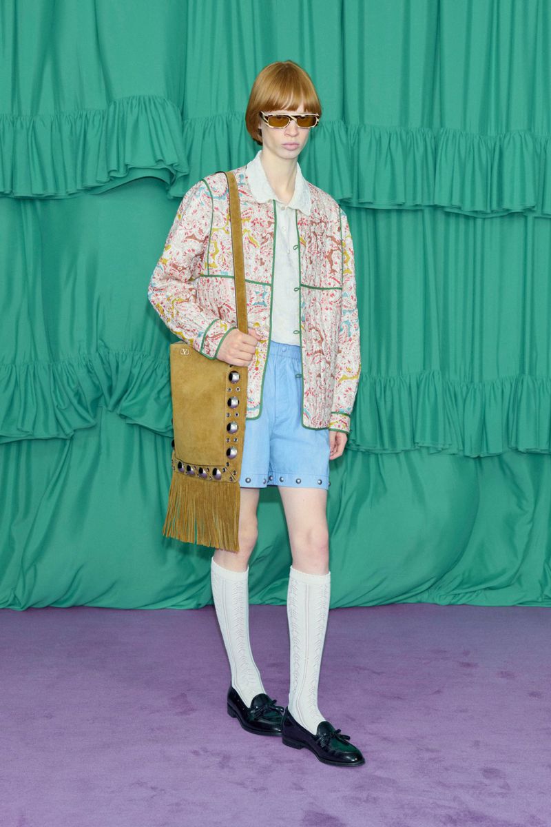 Full-shot fashion portrait: a young adult with red hair, wearing a patterned jacket, shorts, knee-high socks and loafers, posing indoors.