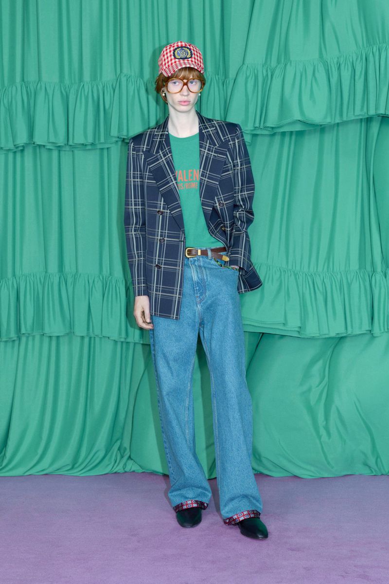 A fair-skinned adult male model wears a plaid blazer and baseball cap in a full-length shot against a layered green curtain backdrop.