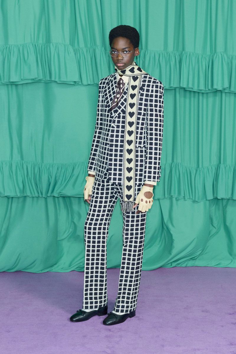 A slender, dark-skinned model poses against a green frilly backdrop in a black and white checkered suit and a heart-patterned scarf.