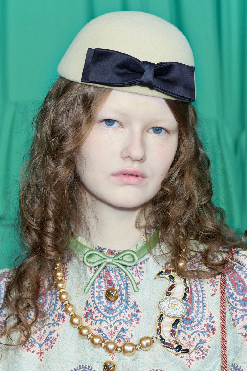 Close-up shot of a woman wearing a light yellow beret with a black bow, a patterned dress, and statement necklaces against a green backdrop.