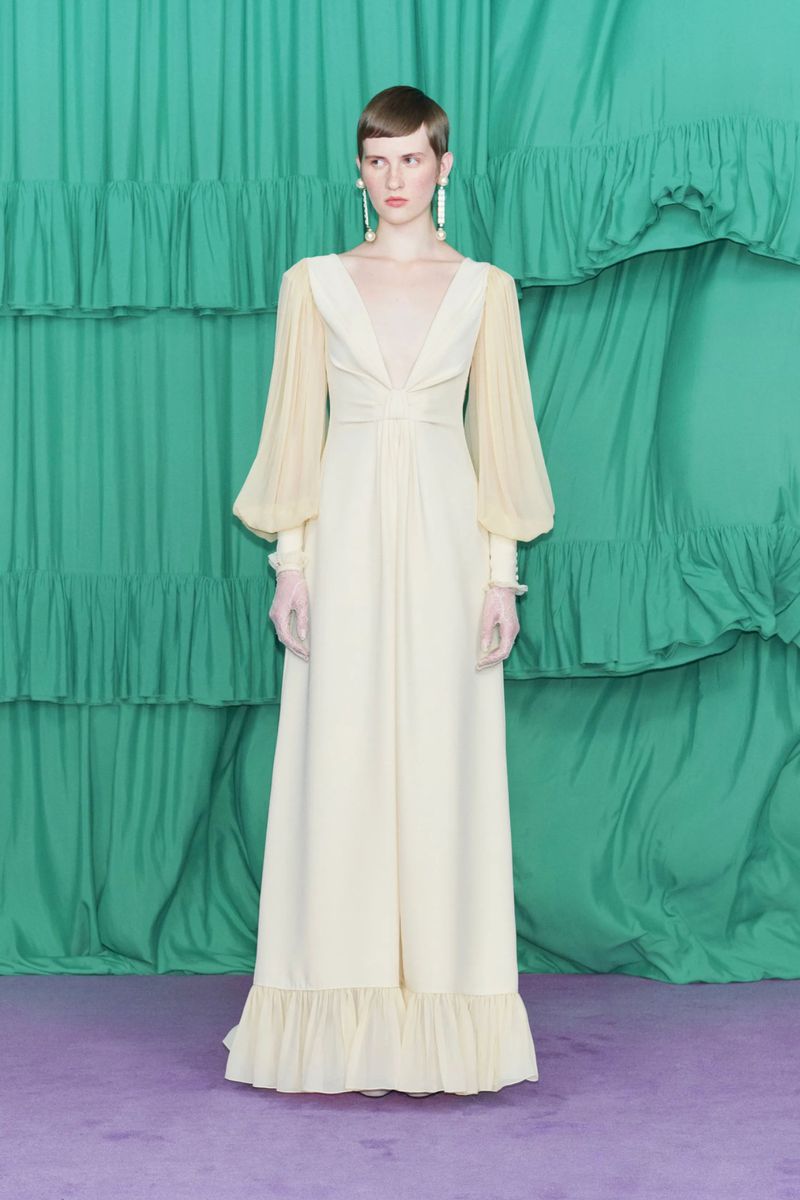 Elegant woman in a long ivory gown with sheer sleeves stands in front of a green ruffled curtain and purple floor.