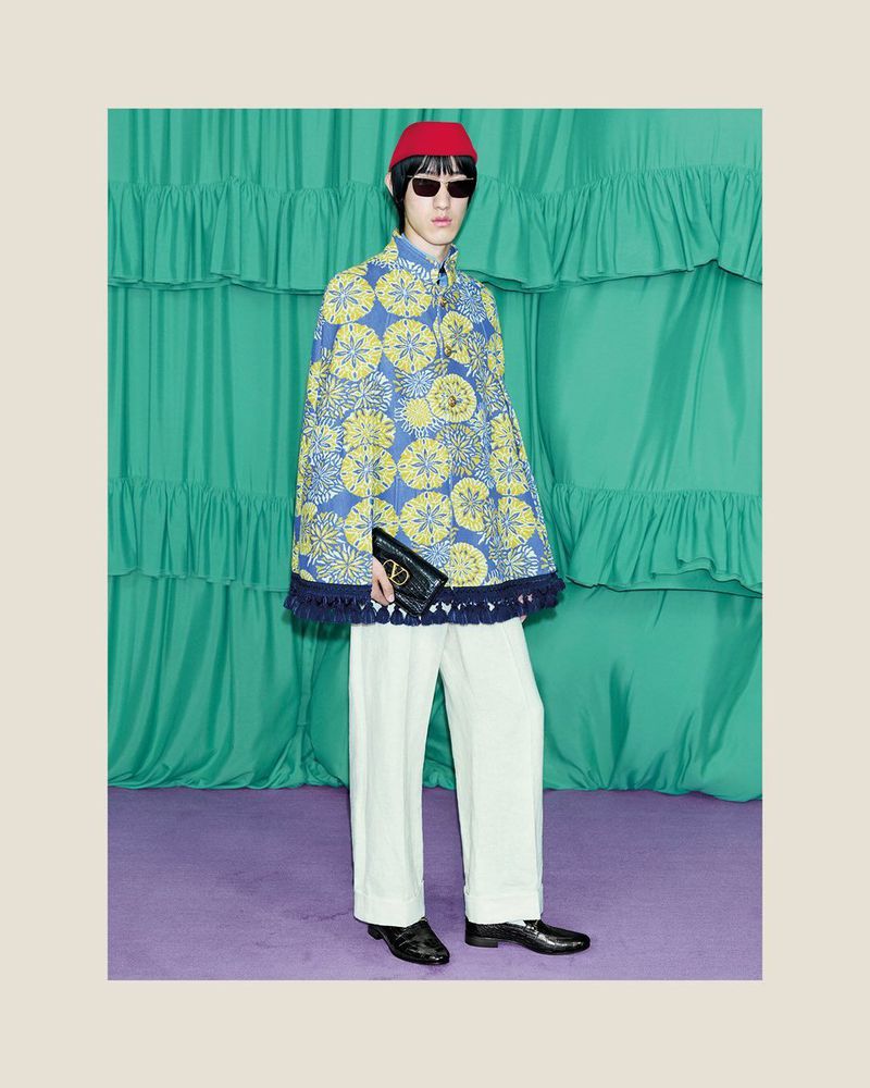 Stylish individual showcasing a blue cape with yellow floral designs, paired with white trousers and black shoes in a studio setting.