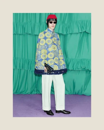 Stylish individual showcasing a blue cape with yellow floral designs, paired with white trousers and black shoes in a studio setting.
