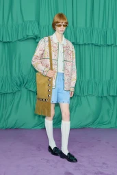 Model wearing a patterned jacket, blue shorts, and a fringed tan suede shoulder bag against a green draped backdrop.