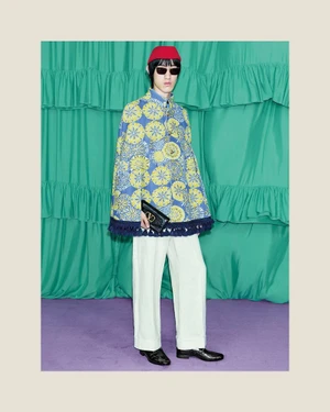 Model wearing a blue patterned cape, white trousers, red cap and sunglasses, holding a black clutch in a green draped studio.