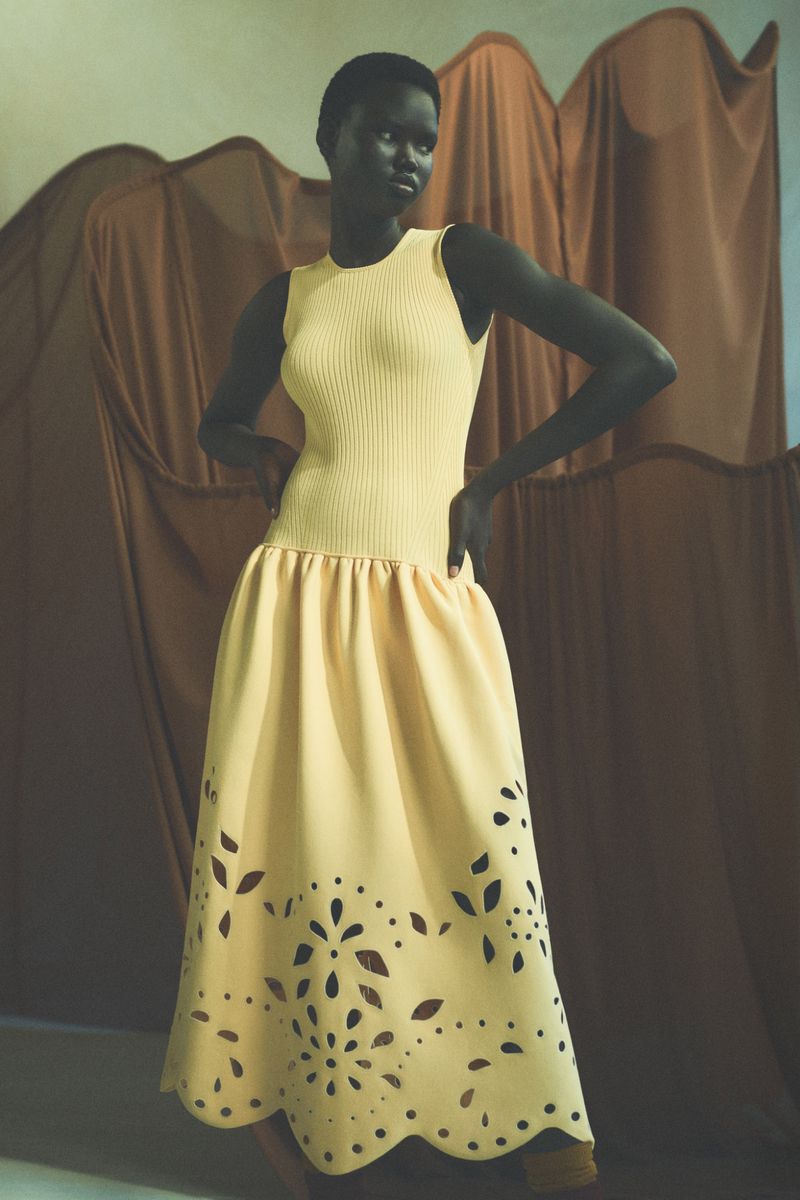 An African American woman poses in a pale yellow ribbed tank top and a matching A-line skirt with cut-out floral designs.