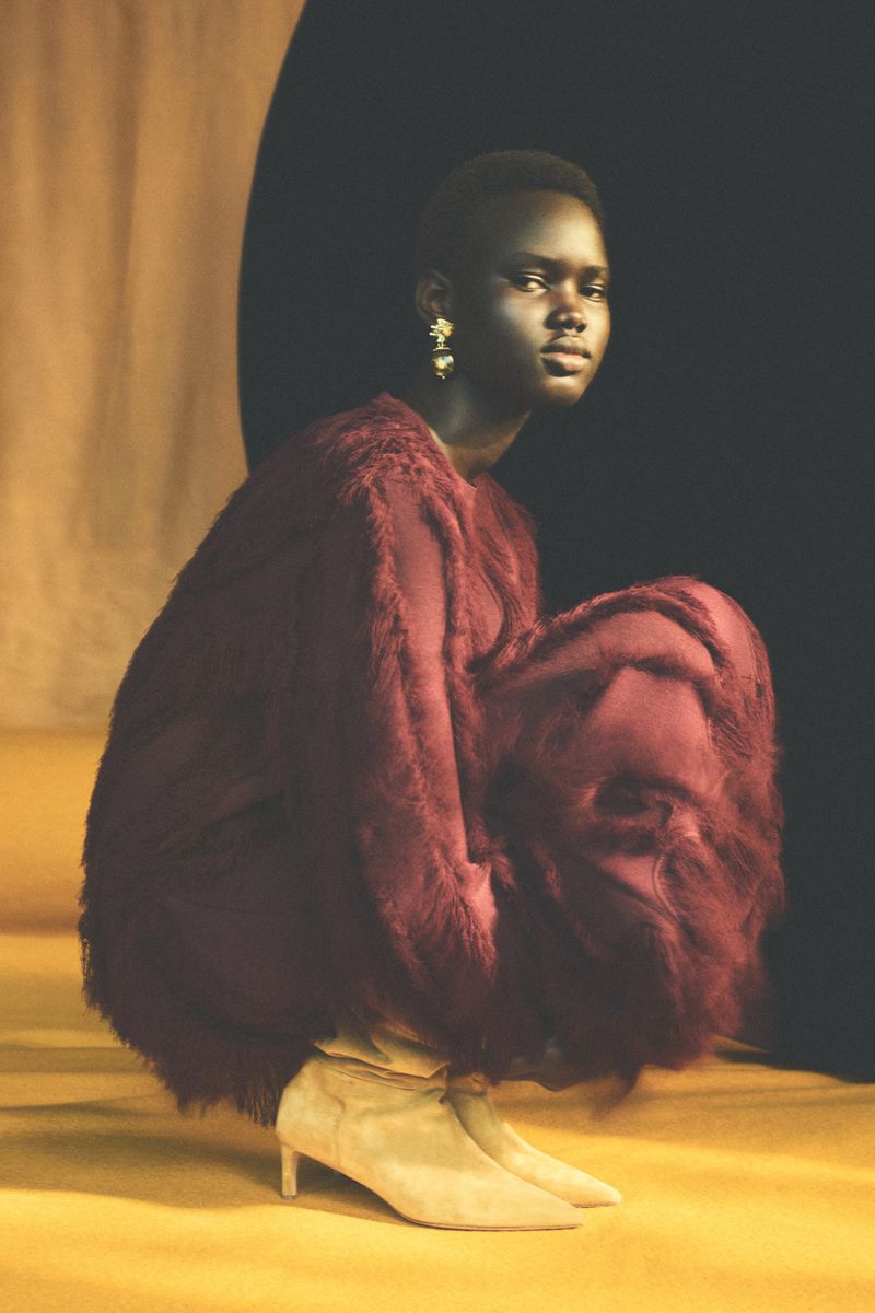 Woman in a red, furry coat crouching on a yellow surface, posing with an elegant gaze. Gold earrings accent the fashion shot.