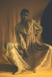 Model wearing a shimmering gold metallic dress with ruffled neckline squatting against a warm textured backdrop
