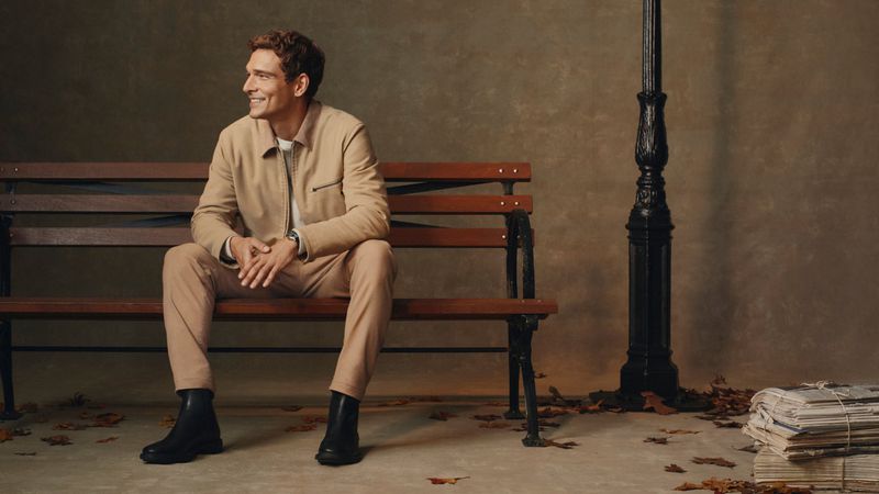 Adult man smiles while seated on a park bench in a beige jacket and pants, with black boots. A black lamppost is behind him.