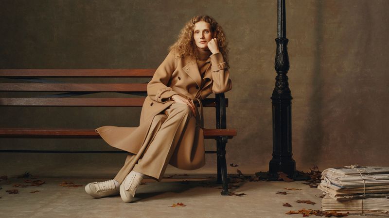 Adult female with wavy brown hair in a tan coat and trousers sits on a brown park bench with a lamp post in the background.
