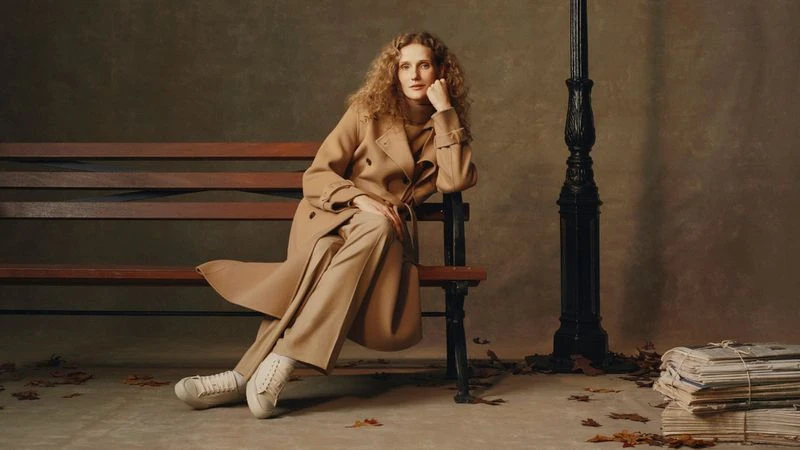 Adult female with wavy brown hair in a tan coat and trousers sits on a brown park bench with a lamp post in the background.