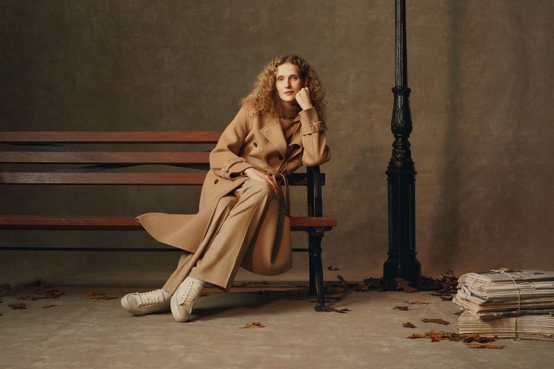 An adult female seated on a park bench beside a lamppost, wearing a tan trench coat and pants, with newspapers on the ground.