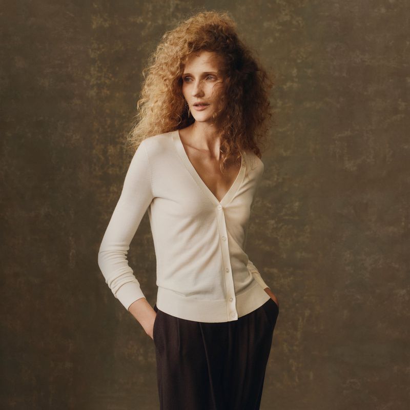 An adult female model poses in a bright photo studio wearing a cream cardigan and dark trousers against a neutral backdrop.