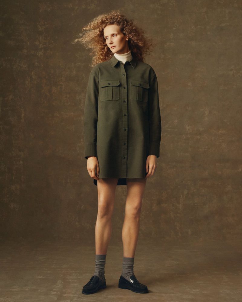 A woman with curly red hair poses in an olive green dress shirt, grey socks, and black loafers against a mottled brown background.