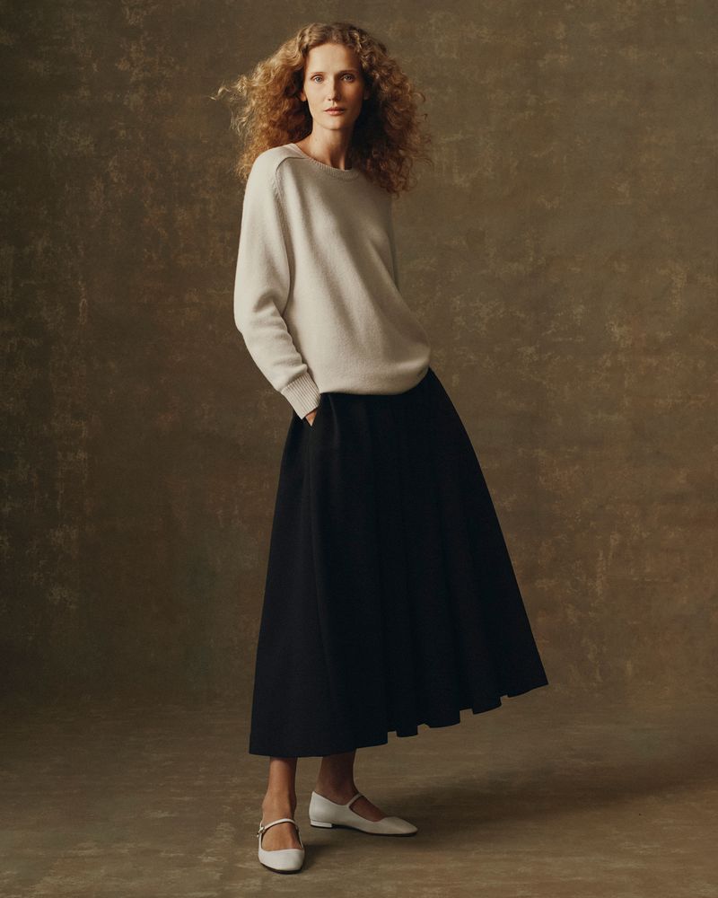 A full-length studio shot of a curly-haired woman in a cream sweater and long black skirt, paired with white Mary Jane shoes, against a tan backdrop.