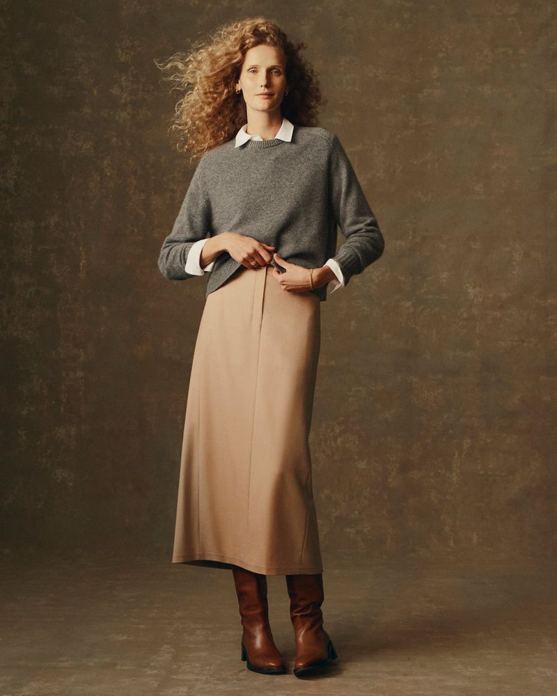 Full-length portrait of a fair-skinned woman in a gray sweater, tan skirt, and brown boots, posing against a mottled brown background.