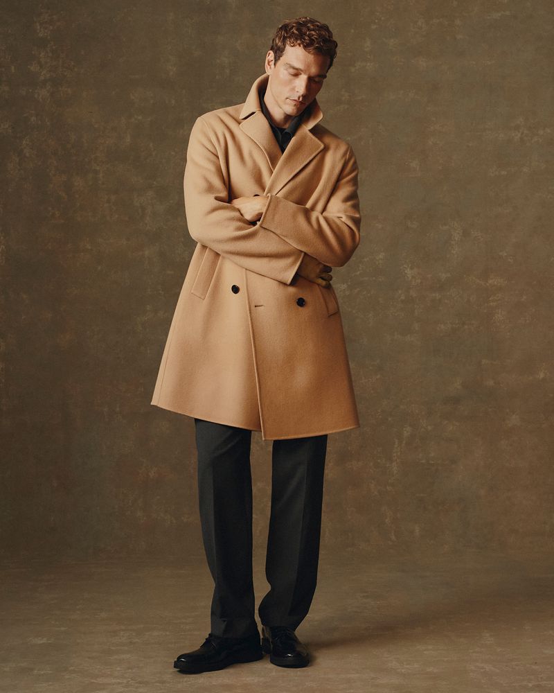 Full-length portrait of an adult male in a camel-colored coat and black dress pants, striking a relaxed pose against a textured backdrop.