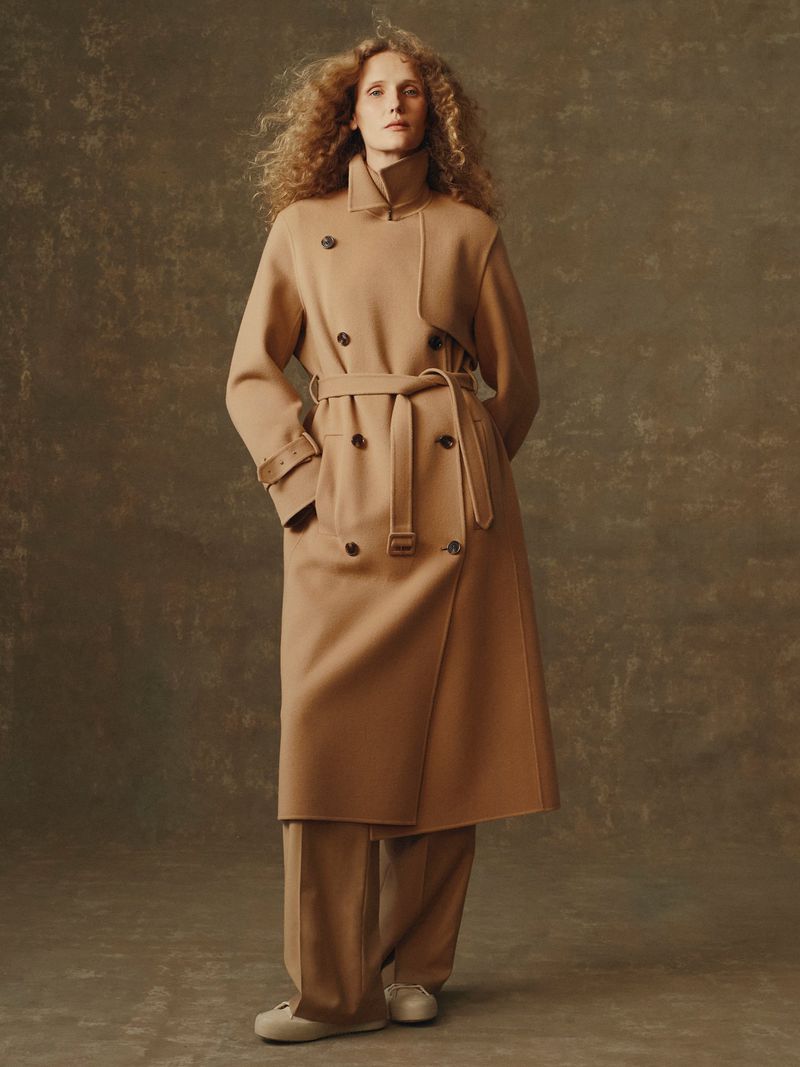 A stylish adult woman wearing a camel colored coat with a tie belt and matching trousers standing indoors in front of a muted background.