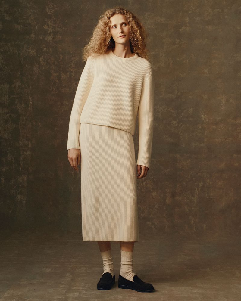 Blonde woman in studio wearing a matching cream-colored ribbed knit sweater and skirt, paired with socks and black loafers.