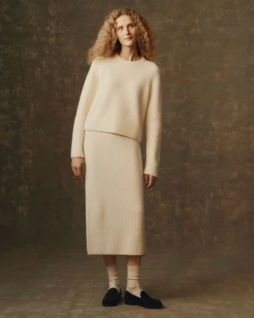 Blonde woman in studio wearing a matching cream-colored ribbed knit sweater and skirt, paired with socks and black loafers.
