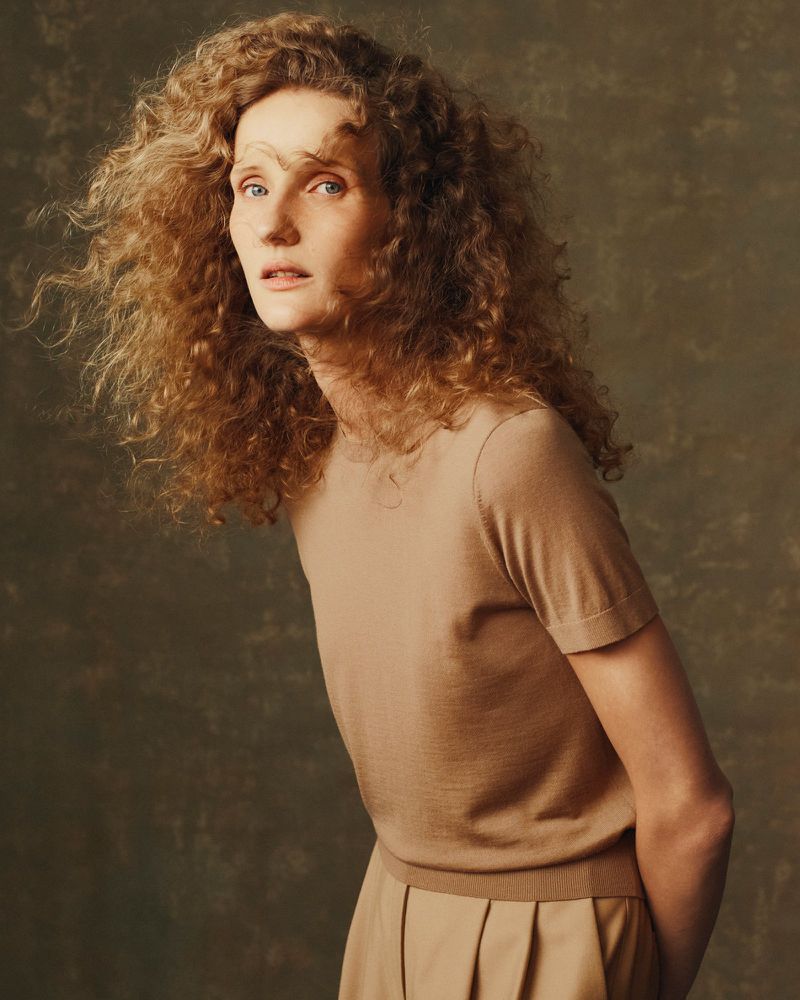 Portrait of a curly-haired woman with fair skin wearing a beige top and skirt against a mottled brown and green background.