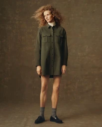 Model with curly blonde hair wearing an olive green tunic shirt and grey socks against a textured backdrop
