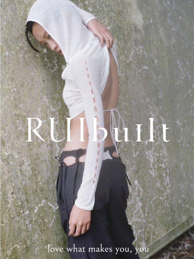 Fashion shot of a slim adult female wearing a white hooded top and black pants, posing against a textured gray wall outdoors.