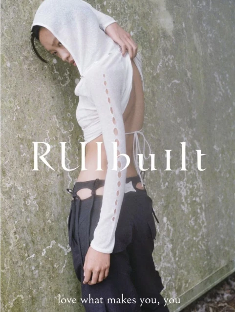 Fashion shot of a slim adult female wearing a white hooded top and black pants, posing against a textured gray wall outdoors.