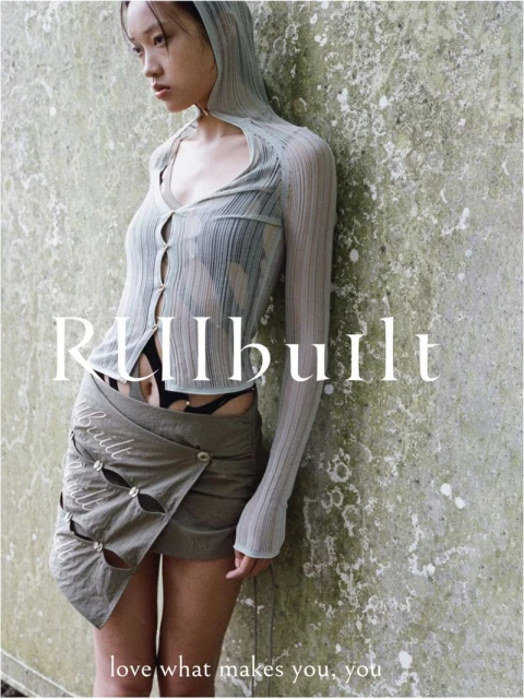 A slender woman in a hooded top and skirt leans against a weathered wall, creating a muted fashion statement in a natural outdoor setting.