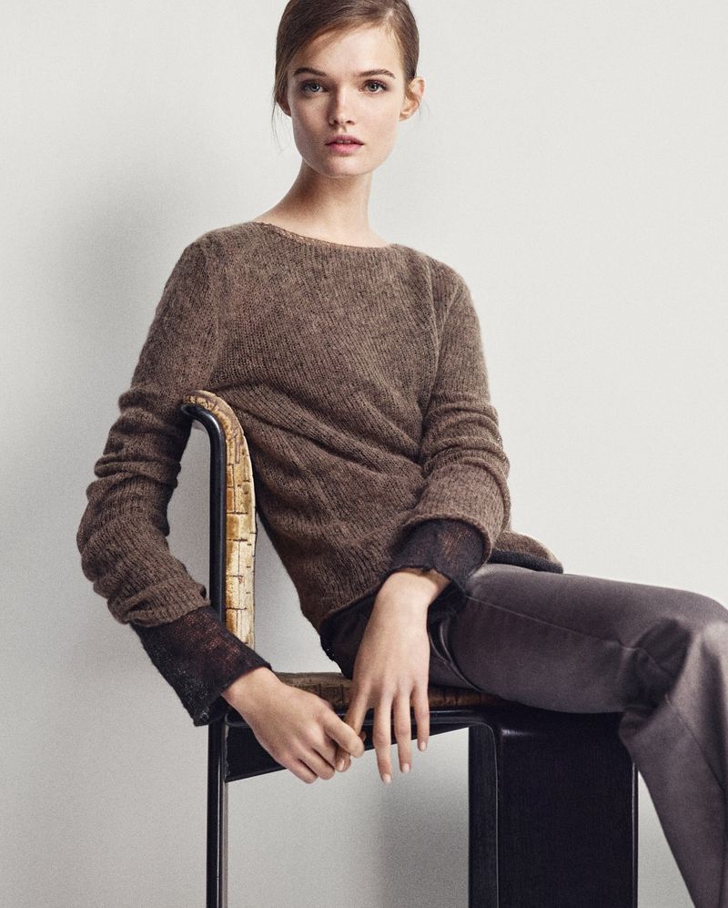 Brunette woman in a brown sweater and grey pants sits in a chair against a white background, posing for a fashion style photograph.