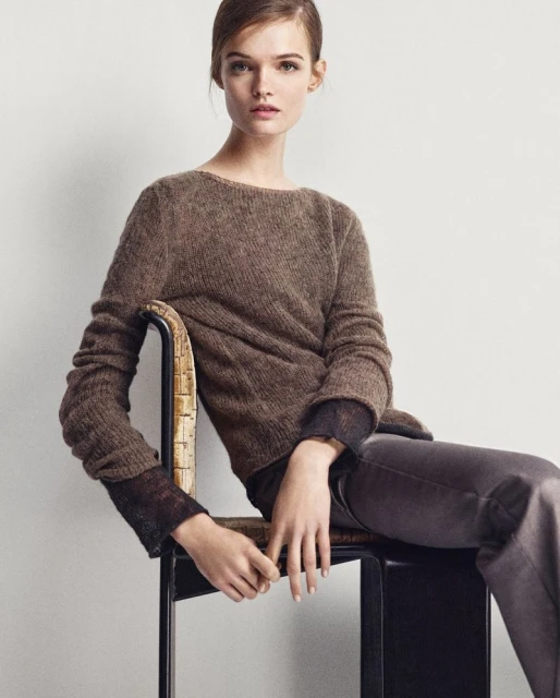 Brunette woman in a brown sweater and grey pants sits in a chair against a white background, posing for a fashion style photograph.