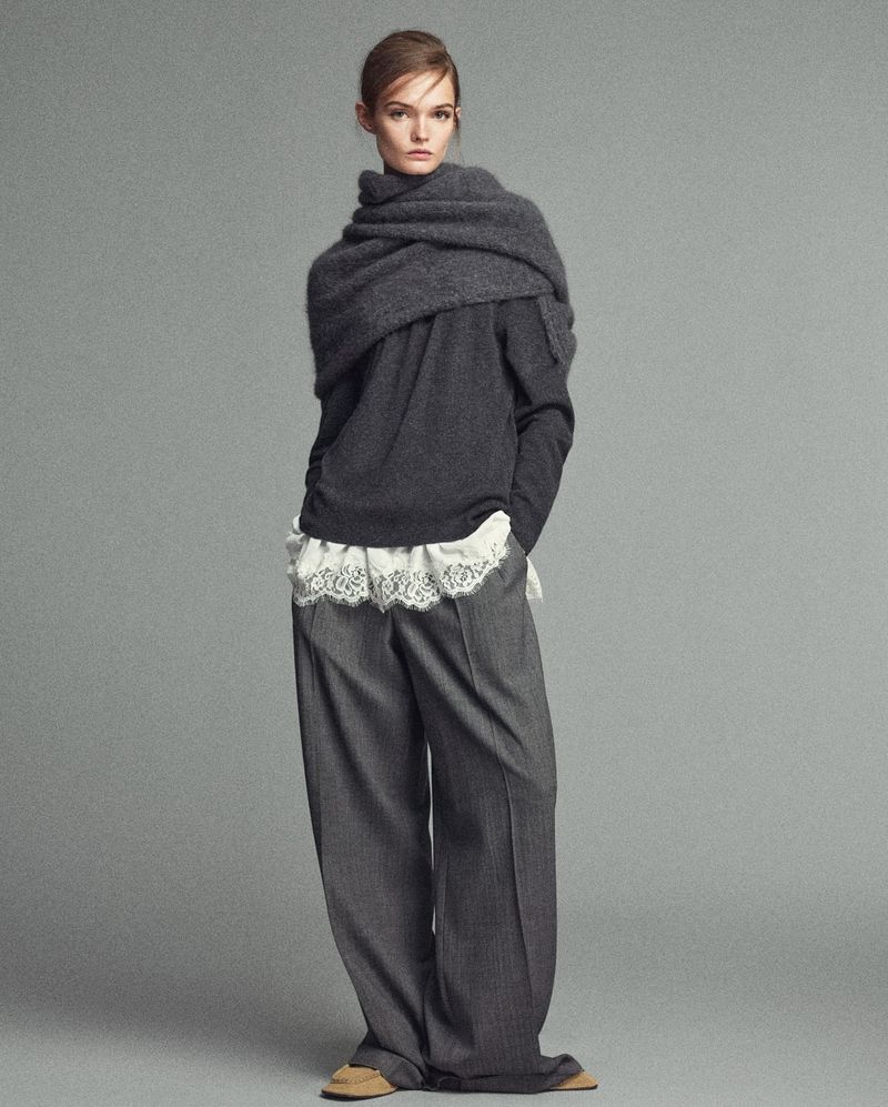 Full-length portrait of a woman in a grey sweater, wide-leg trousers, and a draped scarf on a studio backdrop, showcasing fashion and style.