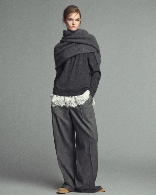 Full-length portrait of a woman in a grey sweater, wide-leg trousers, and a draped scarf on a studio backdrop, showcasing fashion and style.