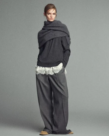Full-length portrait of a woman in a grey sweater, wide-leg trousers, and a draped scarf on a studio backdrop, showcasing fashion and style.
