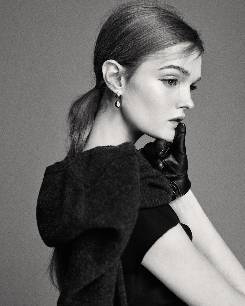 Monochrome portrait of a fair-skinned young woman with brown hair in a ponytail, wearing a black dress, gloves, and statement earrings.