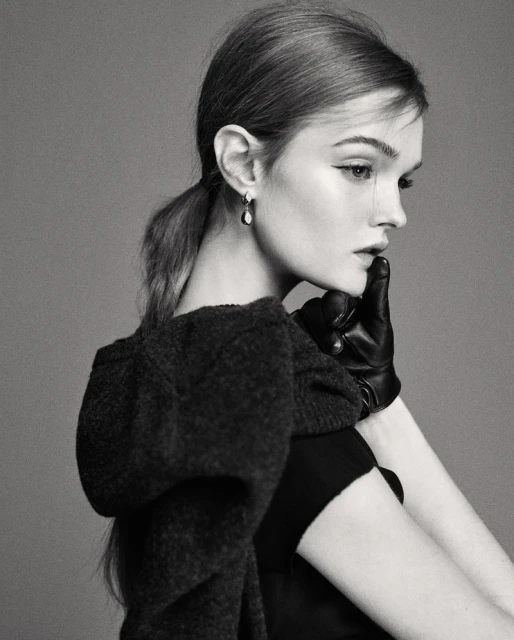 Monochrome portrait of a fair-skinned young woman with brown hair in a ponytail, wearing a black dress, gloves, and statement earrings.